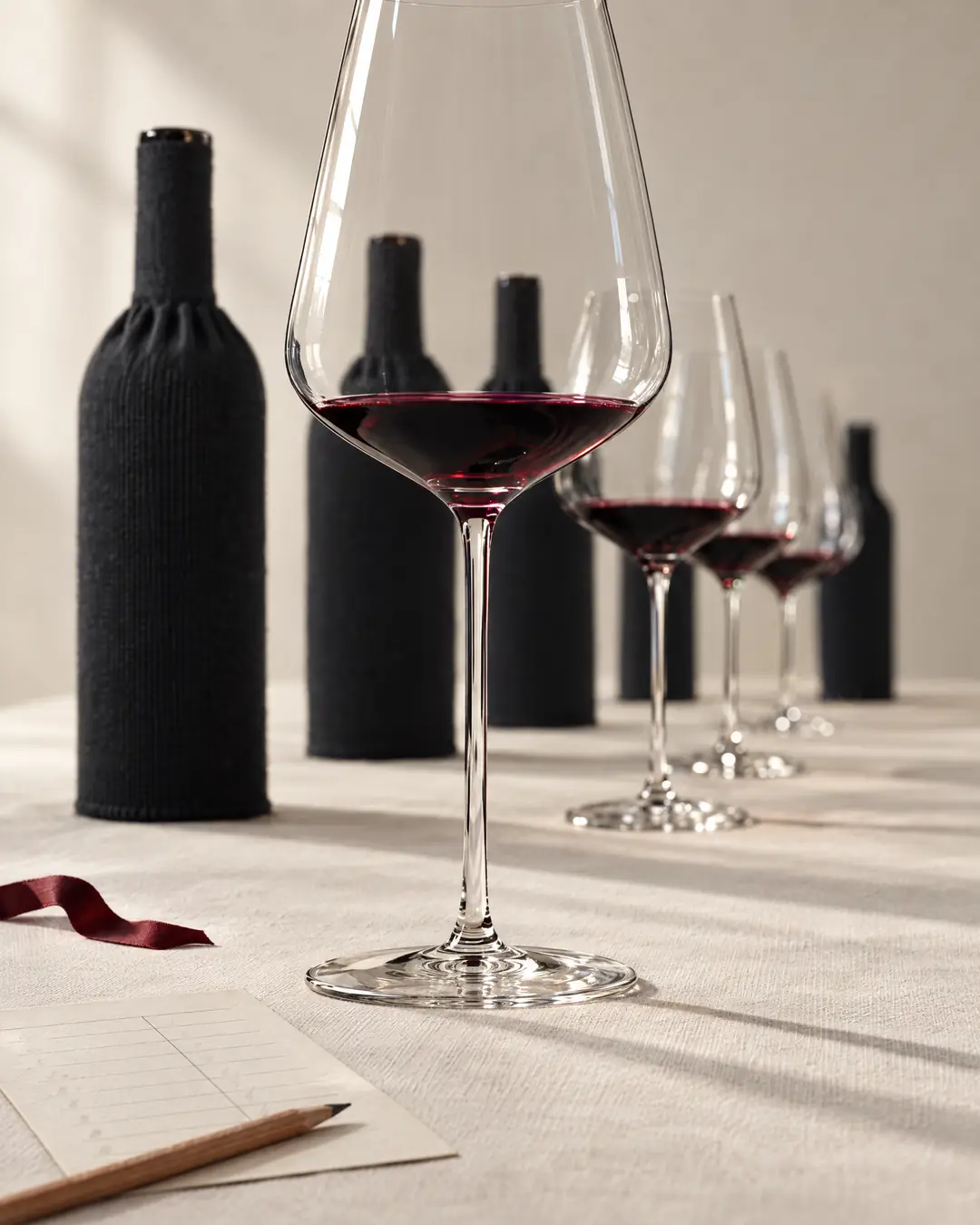 A blind wine tasting under way, a short pour of red in a thin glass before bottles in black knitted sleeves and a ruled scorecard