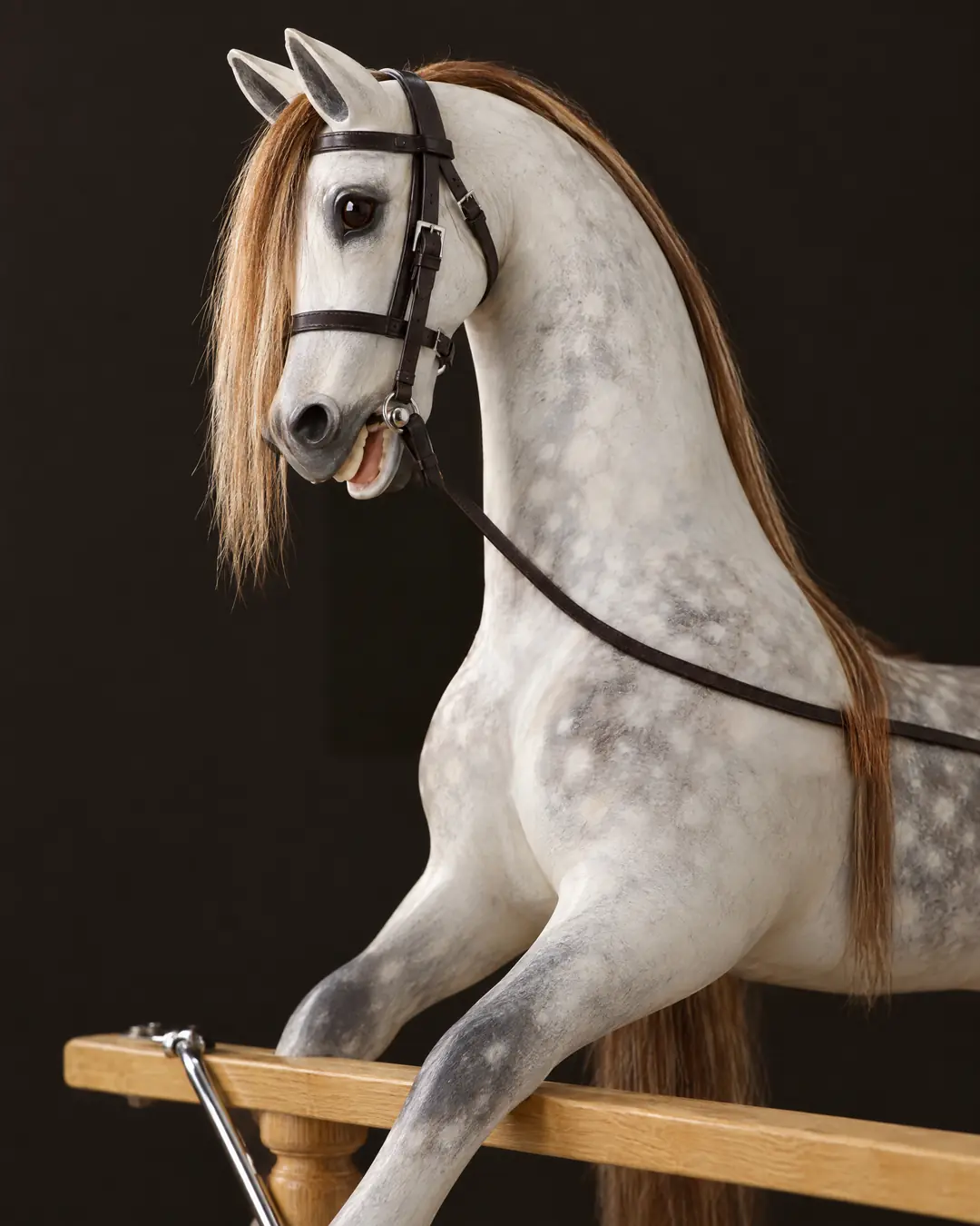 A hand-carved dapple-gray rocking horse with a chestnut horsehair mane and a leather bridle, its forelegs reaching over the oak rail of its stand