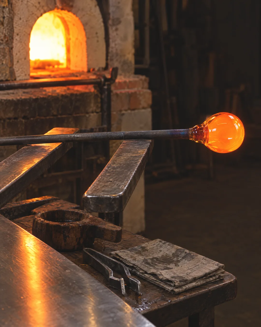 Molten glass glowing orange on the end of a blowpipe at a glassmaker's bench, a Murano furnace glowing behind