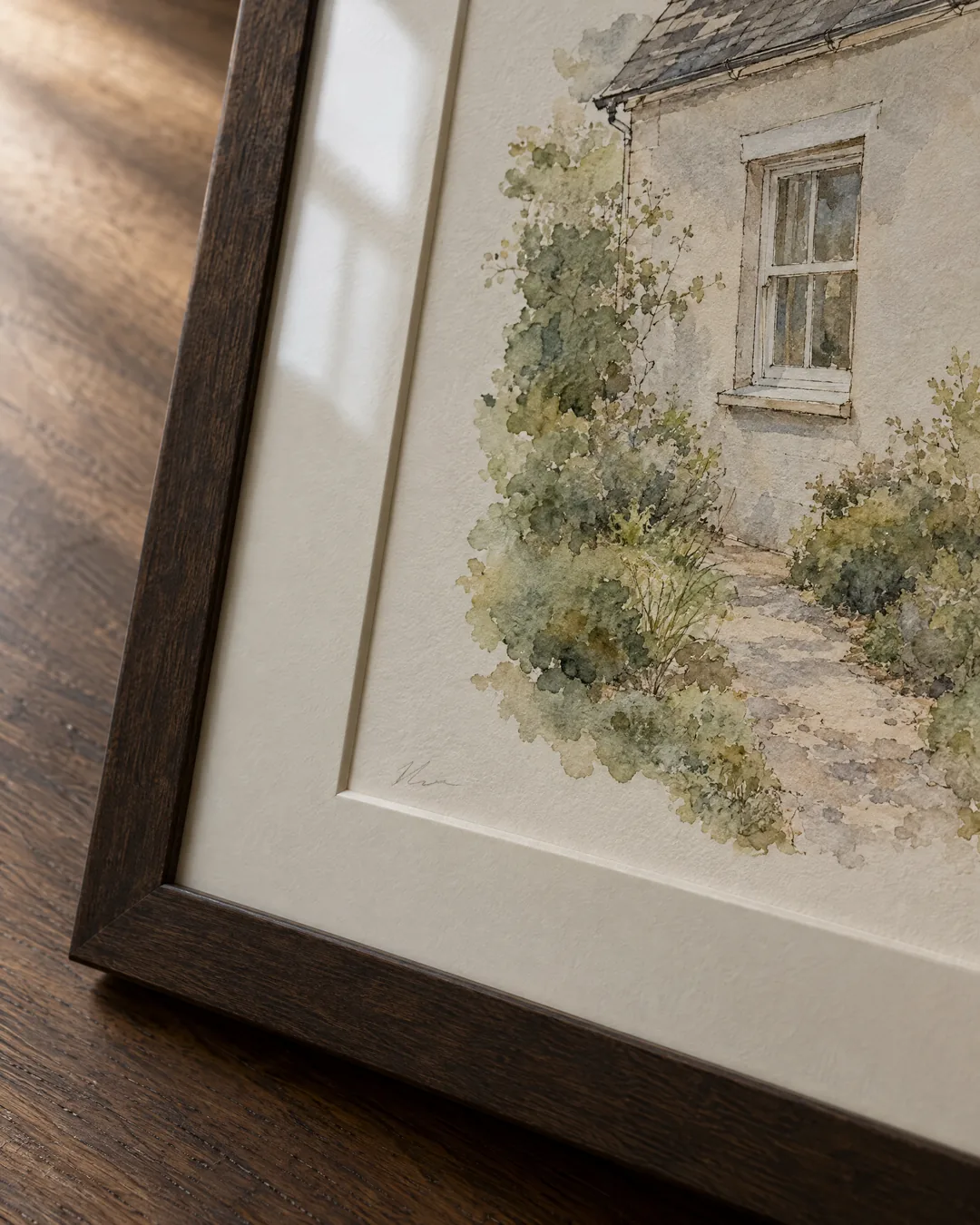 A framed watercolor of the corner of a family house, with a sash window and a garden path, leaning on a dark oak table