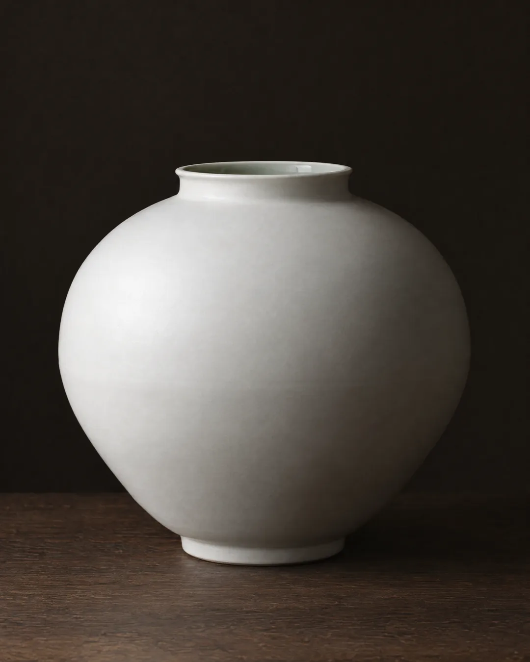 A white porcelain moon jar with a matt glaze and a faint join at its widest point, on a dark oak table