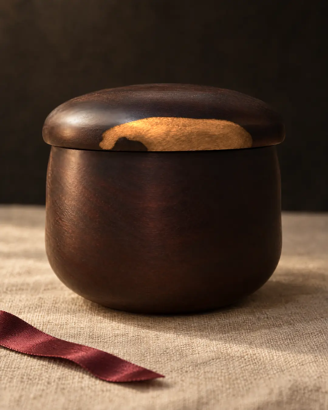 A small, nearly black wooden bowl with a domed lid and a streak of golden sapwood along the lid's edge, on linen in low side light, a dark red ribbon beside it