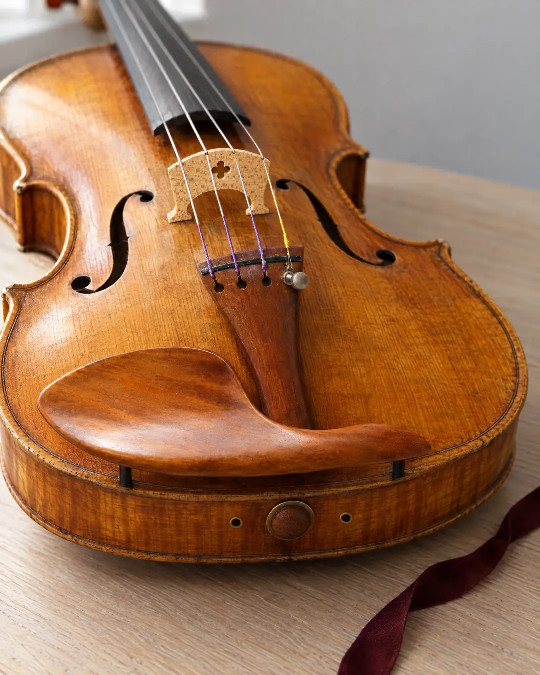 A golden-orange violin seen close from its tail end on pale oak, a carved wooden chinrest arching over the tailpiece and four strings running over a pale maple bridge, with an oxblood ribbon beside it