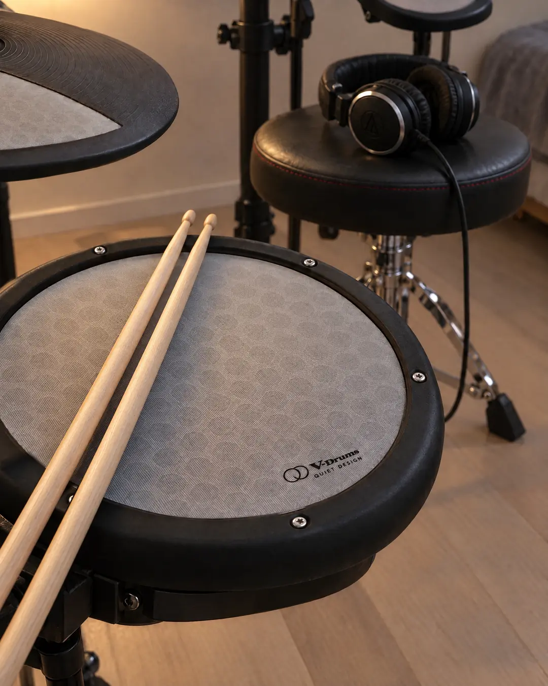 Two hickory drumsticks resting across the pale mesh snare of a compact electronic drum kit, with a cymbal pad nearby and headphones waiting on the drum throne, in soft evening lamplight