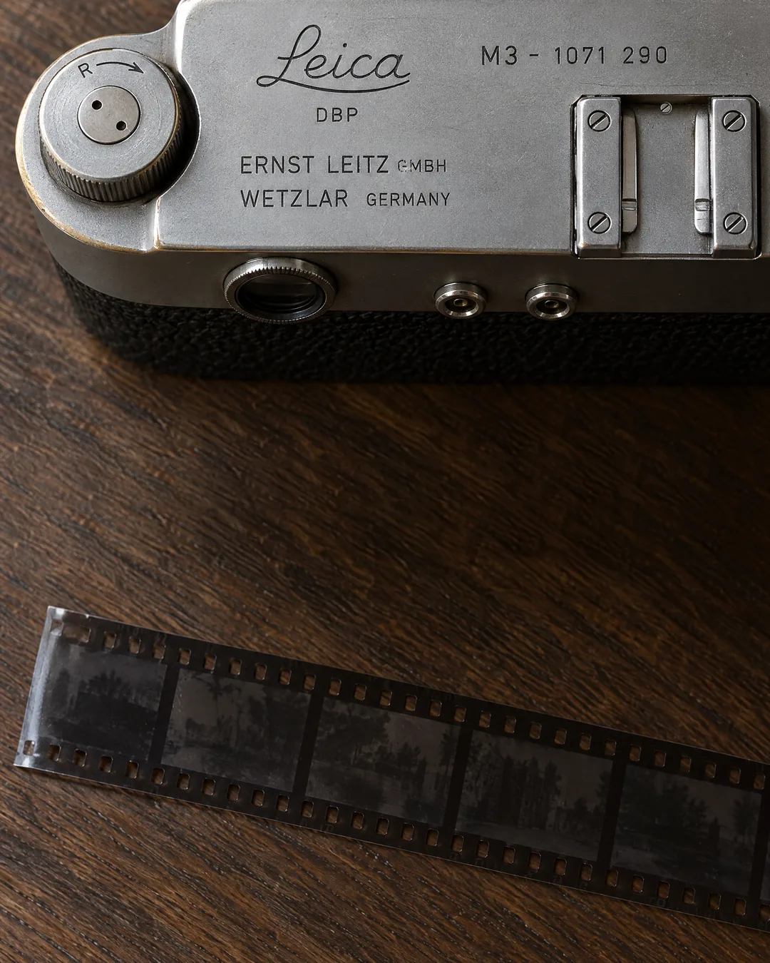 The chrome top plate of a Leica M3 rangefinder seen from above, engraved Ernst Leitz GmbH Wetzlar Germany and serial 1071 290 beside its rewind knob, with a strip of black-and-white negatives on dark oak