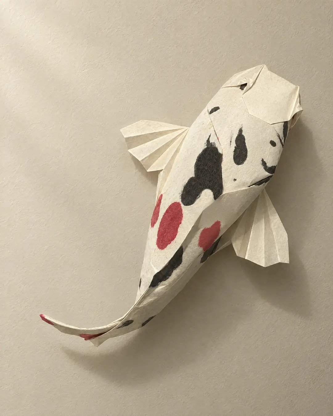 An origami koi folded from one sheet of ivory handmade paper with black and crimson markings in the paper, its pleated fins spread, casting a soft shadow on a pale plaster wall