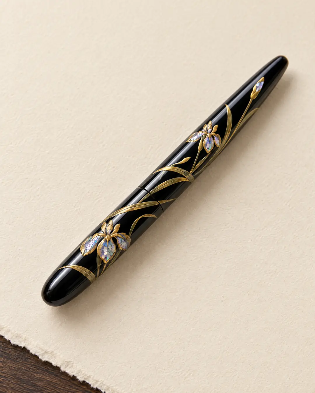 A black lacquer fountain pen painted with raised gold irises, mother-of-pearl in the petals, lying capped on cream paper