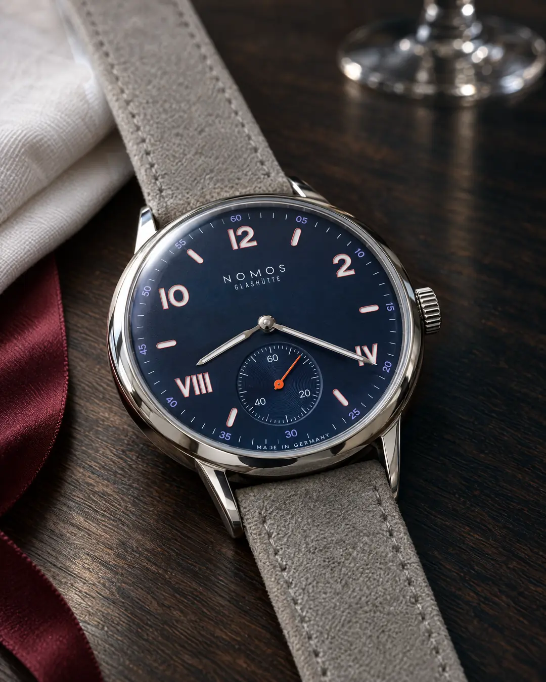 A small NOMOS steel watch with a navy dial, white numerals and an orange seconds hand on a gray strap, lying on a dark oak dining table beside a folded white napkin