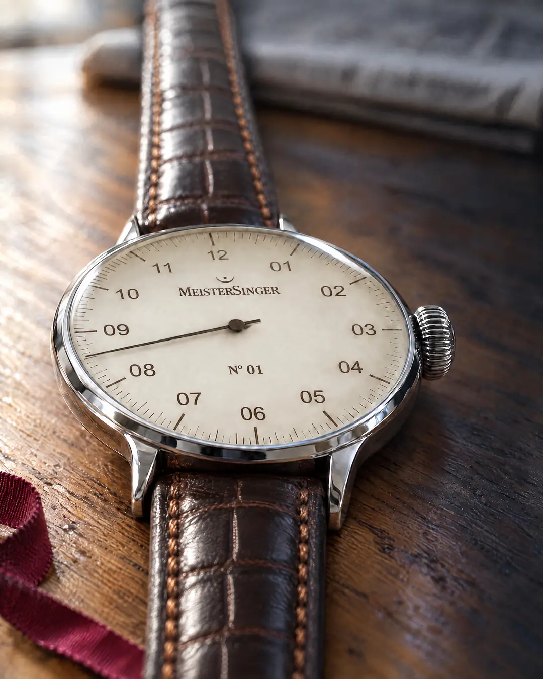 A single-hand steel watch with an ivory dial and brown leather strap on a kitchen table, a folded morning newspaper behind it